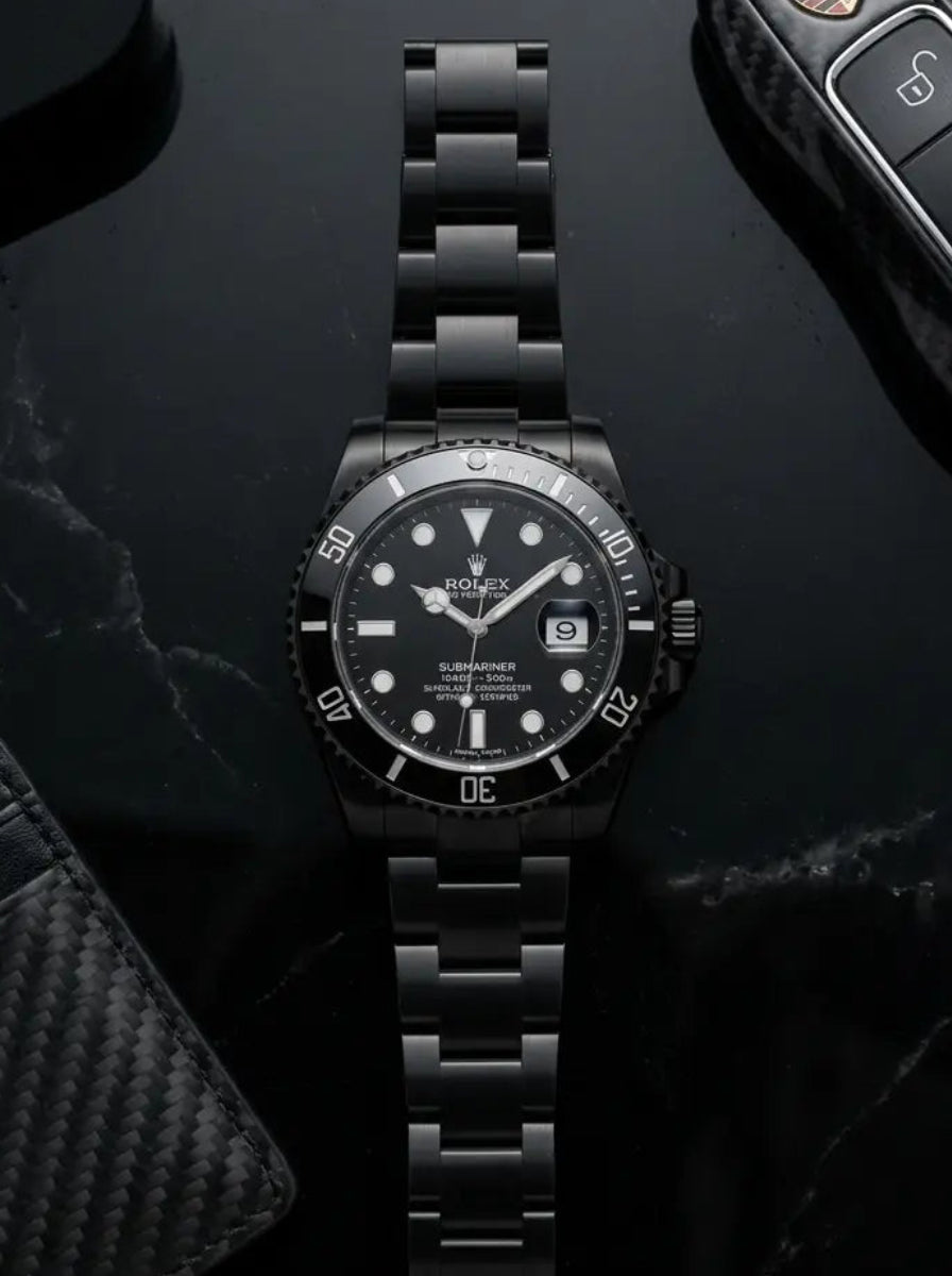 Black watch on a dark surface with a car key fob and wallet.